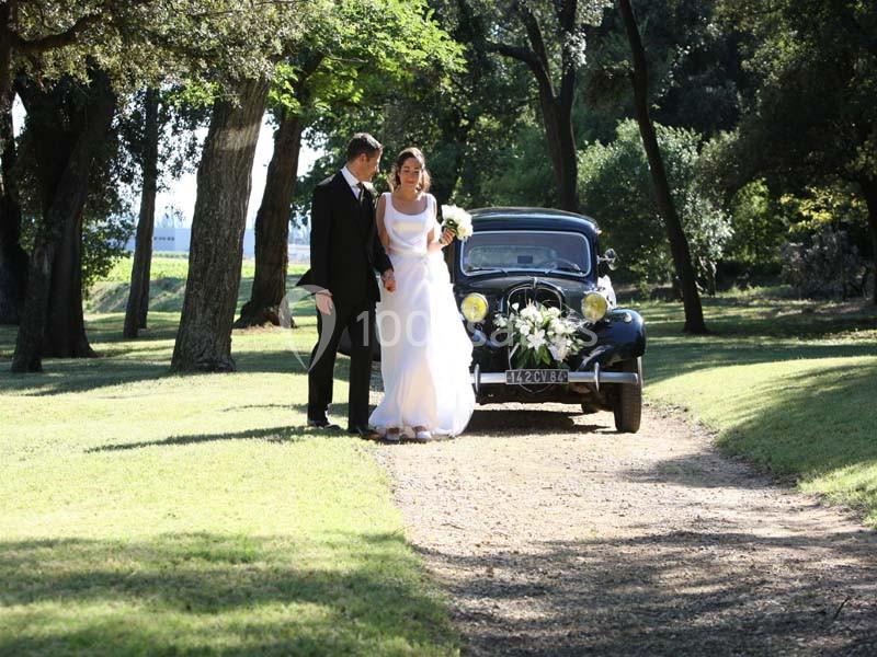 Location salle Piolenc (Vaucluse) - Château Beauchêne #16 Un couple en tenue de mariage marche sur un chemin de terre, avec une voiture ancienne décorée en arrière-plan.
