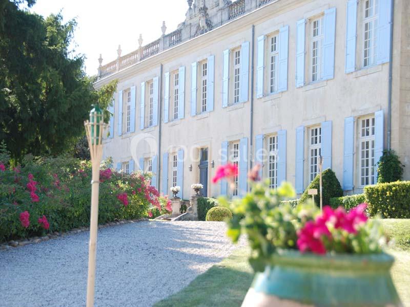 Location salle Piolenc (Vaucluse) - Château Beauchêne #4 Façade d'un grand bâtiment ancien avec volets bleus, jardin fleuri et allée gravillonnée au premier plan.