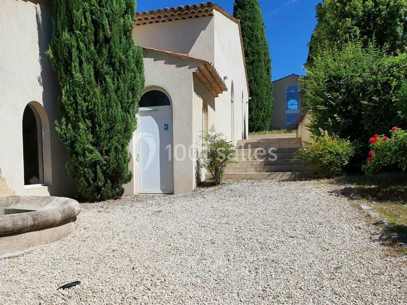 Cour extérieure avec gravier, escalier en pierre, façade beige, portes blanches, cyprès et buissons fleuris.