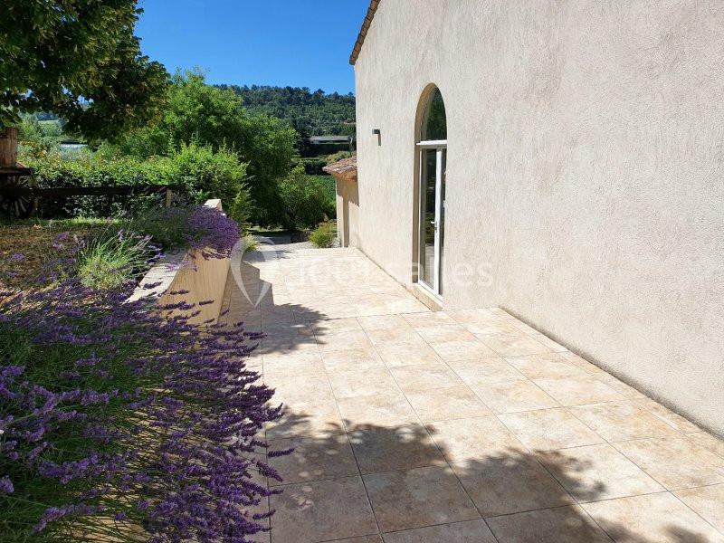 Terrasse en carrelage clair bordée de lavandes, avec vue sur un jardin verdoyant et des collines en arrière-plan.