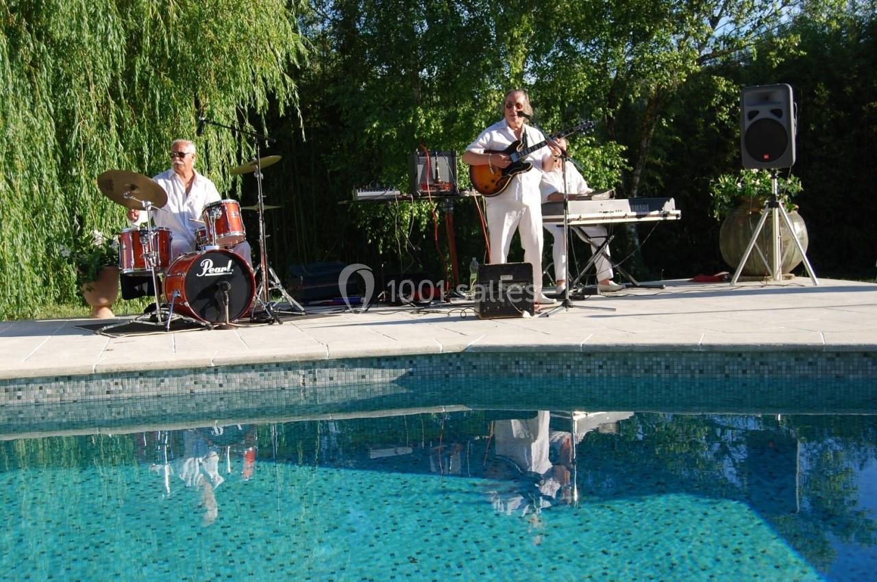 Deux musiciens jouent près d'une piscine, l'un à la batterie et l'autre à la guitare, entourés de verdure.