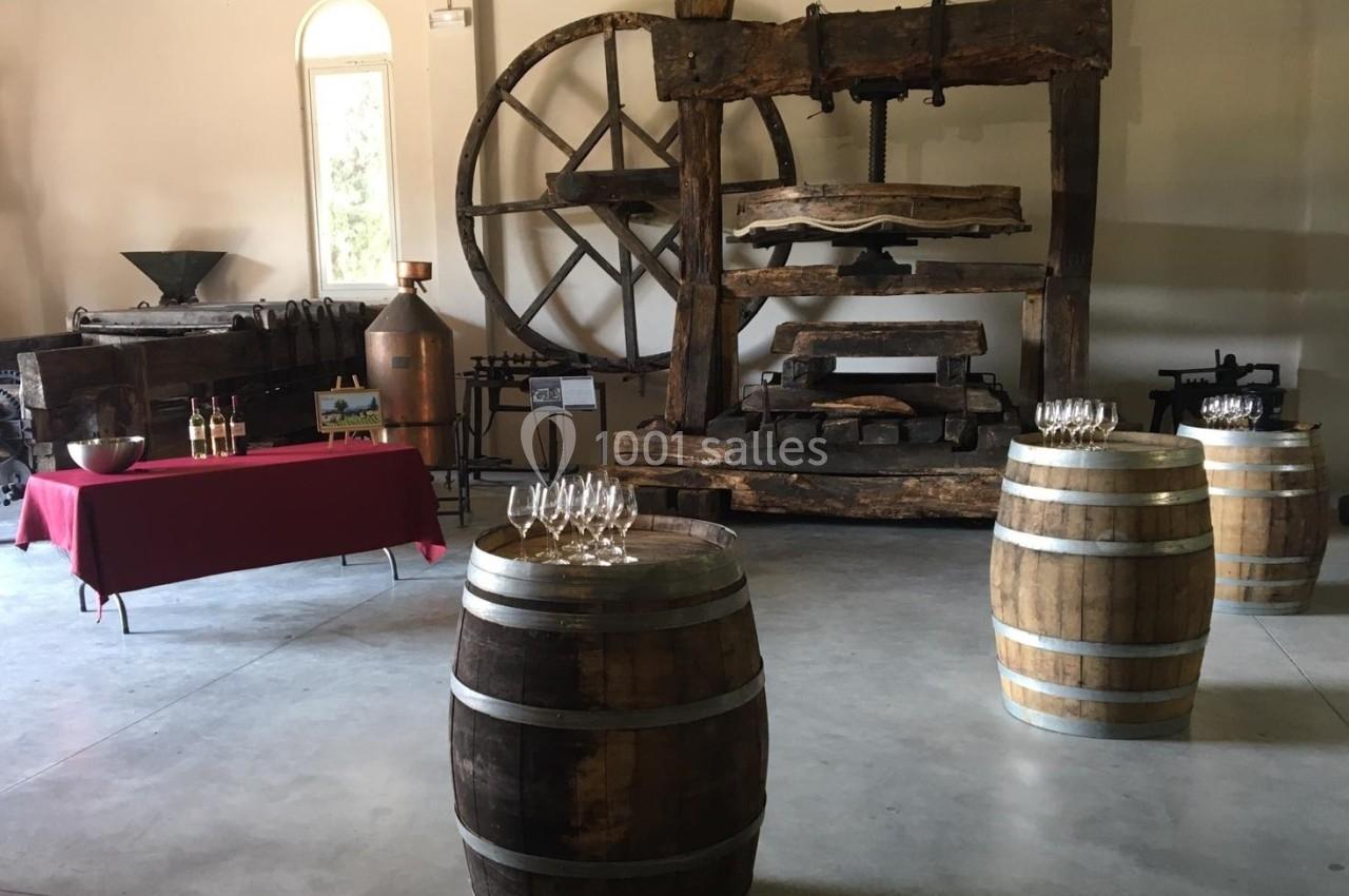 Salle avec un ancien pressoir en bois, des tonneaux, une table rouge avec bouteilles et verres, et des outils viticoles.