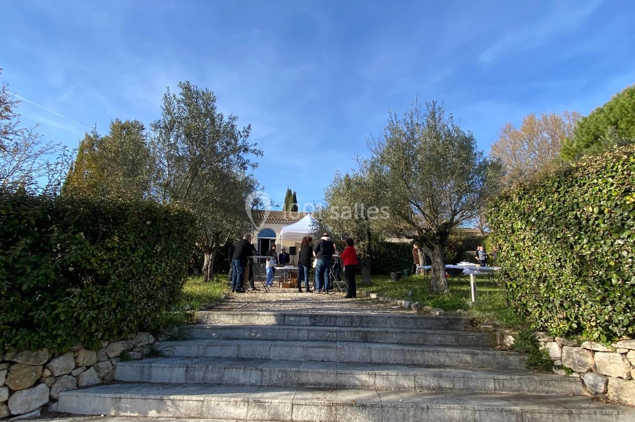 Groupe de personnes rassemblées dans un jardin devant une maison, entouré d'arbres et de haies sous un ciel bleu.