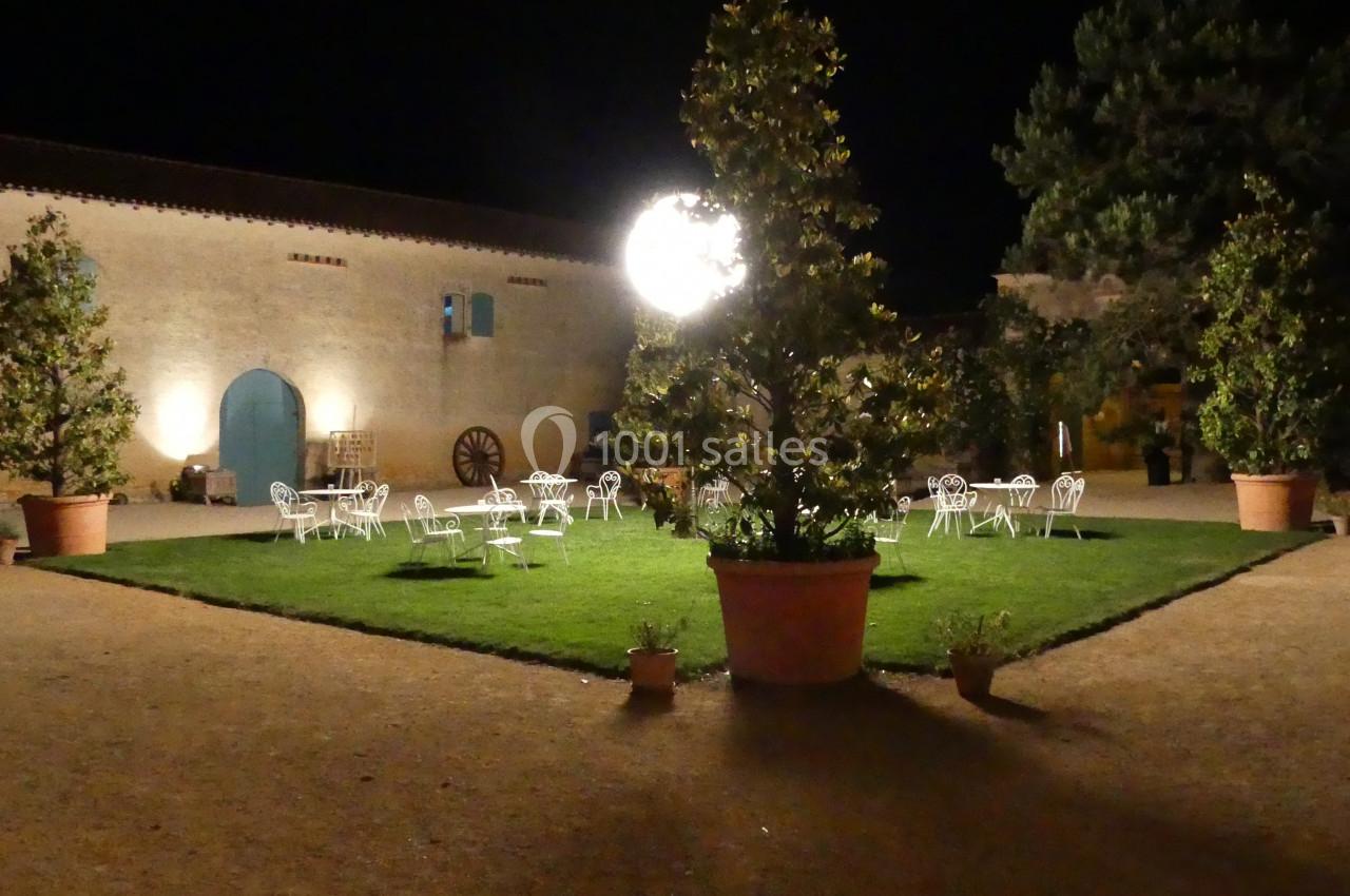 Location salle Sainte-Gemme-la-Plaine (Vendée) - Château de la Chevallerie #24 Cour extérieure éclairée la nuit, avec des tables et chaises blanches disposées autour d'une pelouse centrale.