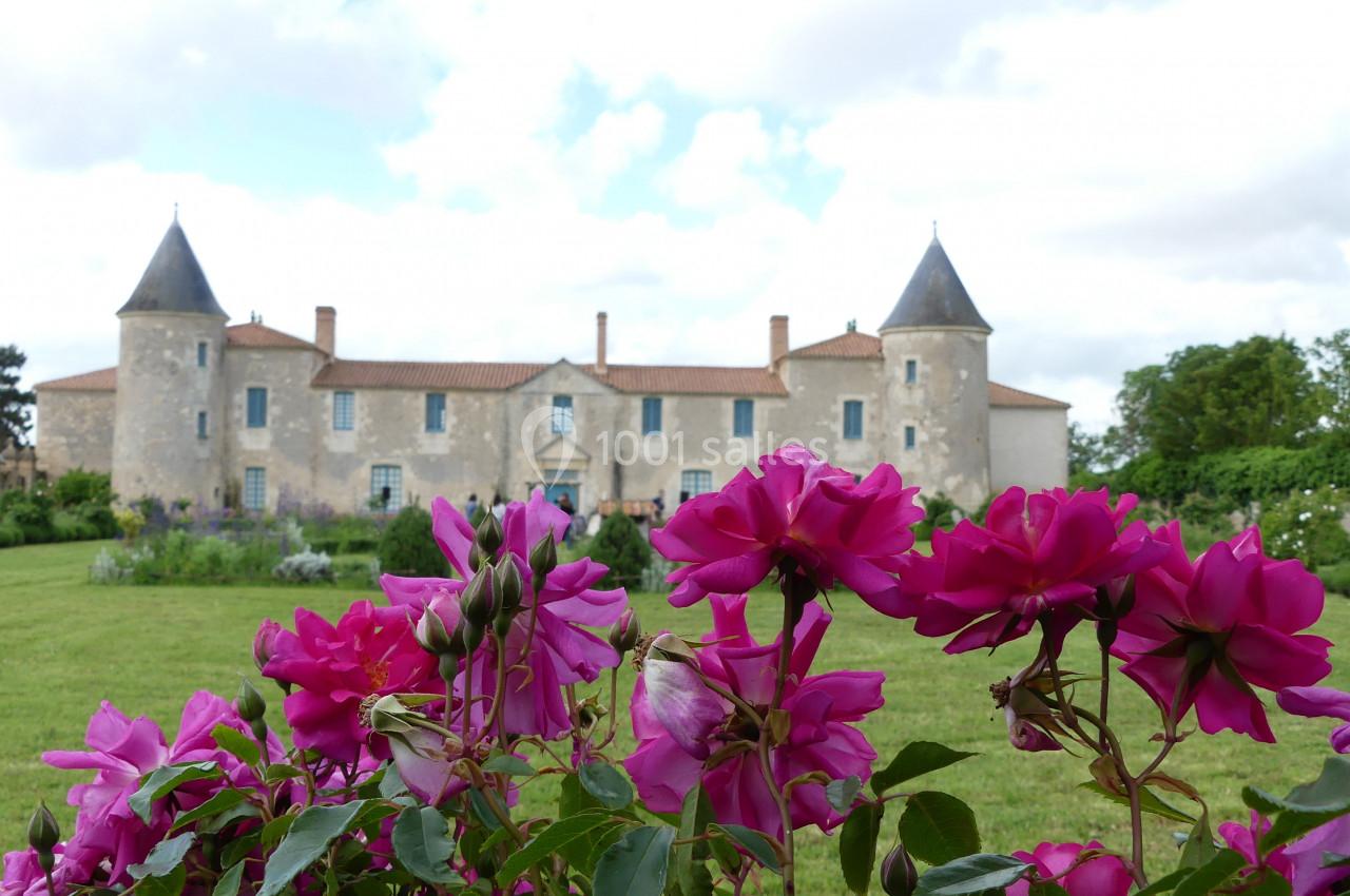 Location salle Sainte-Gemme-la-Plaine (Vendée) - Château de la Chevallerie #16 Château en pierre avec tours, entouré d'un jardin verdoyant, vu à travers des roses roses au premier plan.