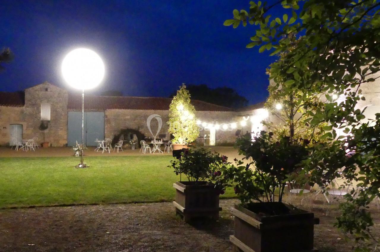 Location salle Sainte-Gemme-la-Plaine (Vendée) - Château de la Chevallerie #26 Cour extérieure éclairée de nuit avec des guirlandes lumineuses, des tables et des plantes en pot.