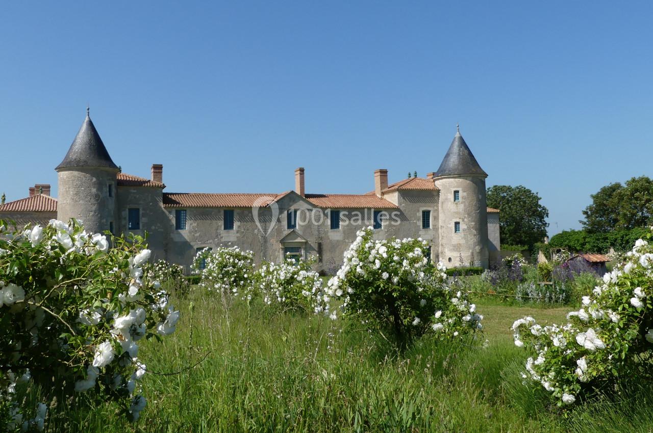 Location salle Sainte-Gemme-la-Plaine (Vendée) - Château de la Chevallerie #14 Château en pierre avec deux tours, entouré de rosiers blancs et d'un jardin verdoyant sous un ciel bleu.