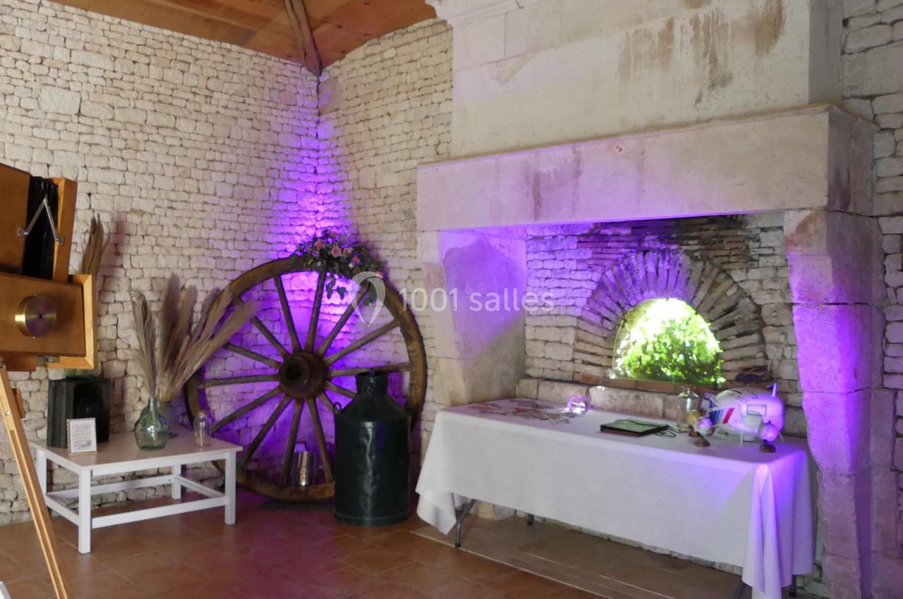 Location salle Sainte-Gemme-la-Plaine (Vendée) - Château de la Chevallerie #32 Salle en pierre avec une cheminée, une roue en bois décorative, une table blanche et un éclairage violet.