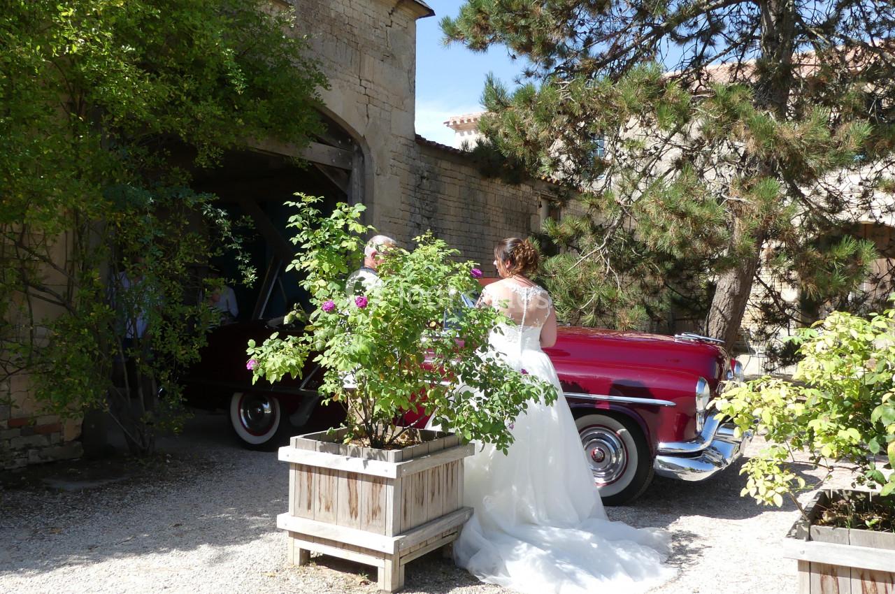 Location salle Sainte-Gemme-la-Plaine (Vendée) - Château de la Chevallerie #35 Une mariée en robe blanche se tient près d'une voiture rouge ancienne, entourée de verdure et de bâtiments en pierre.