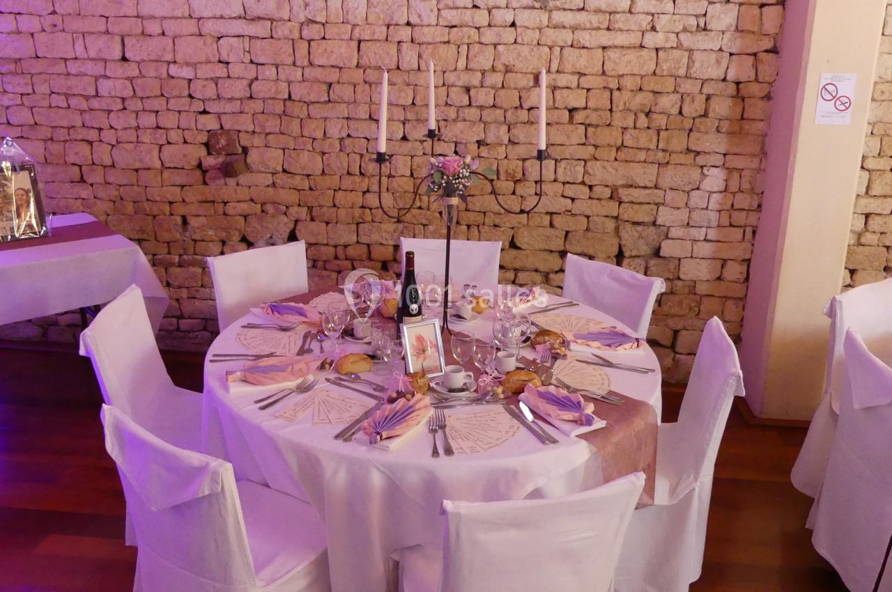 Location salle Sainte-Gemme-la-Plaine (Vendée) - Château de la Chevallerie #22 Table ronde décorée pour un événement, avec nappes blanches, chandeliers, menus et couverts, devant un mur en pierre.