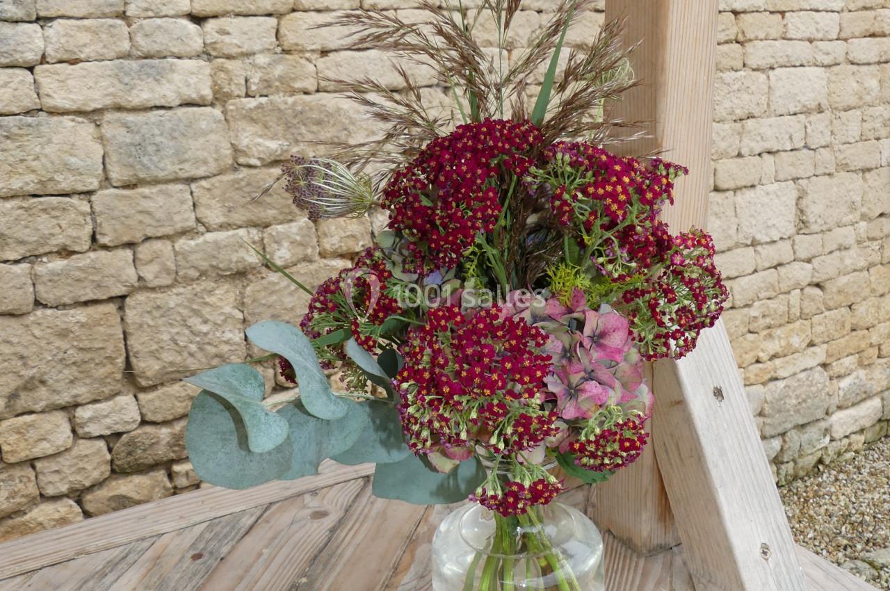 Location salle Sainte-Gemme-la-Plaine (Vendée) - Château de la Chevallerie #37 Bouquet de fleurs rouges et roses avec des feuillages dans un vase en verre, posé sur une table en bois devant un mur en…