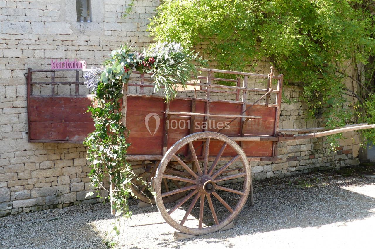 Location salle Sainte-Gemme-la-Plaine (Vendée) - Château de la Chevallerie #40 Charrette en bois décorée de fleurs et feuillages, adossée à un mur en pierre dans un espace extérieur ensoleillé.