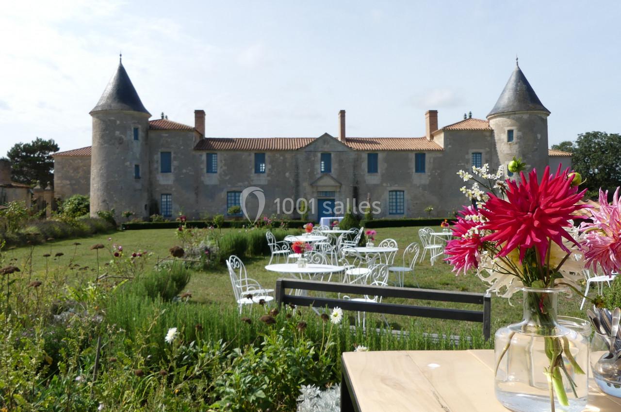 Location salle Sainte-Gemme-la-Plaine (Vendée) - Château de la Chevallerie #2 Façade d'un château avec tours, jardin fleuri, mobilier de jardin blanc et vase de fleurs sur une table au premier plan.