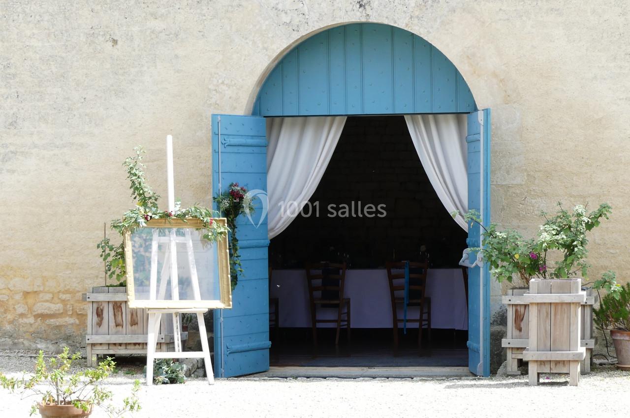 Location salle Sainte-Gemme-la-Plaine (Vendée) - Château de la Chevallerie #41 Porte bleue ouverte sur une salle avec des chaises, encadrée de plantes en pots et de rideaux blancs.