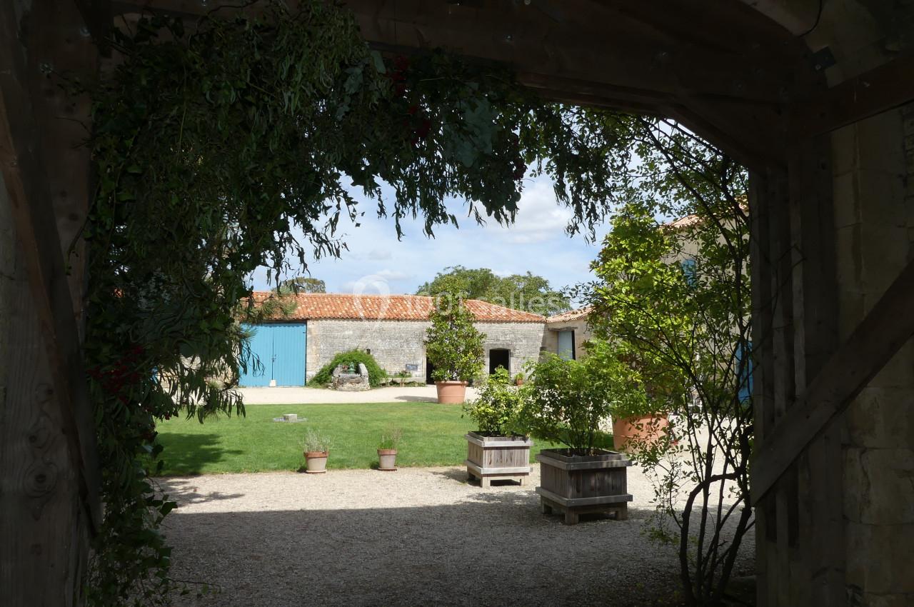 Location salle Sainte-Gemme-la-Plaine (Vendée) - Château de la Chevallerie #42 Vue d'une cour intérieure avec bâtiments en pierre, toits en tuiles, pelouse et jardinières sous une arche en bois.