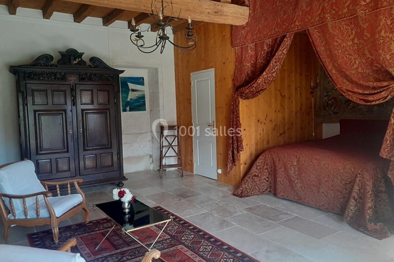 Location salle Sainte-Gemme-la-Plaine (Vendée) - Château de la Chevallerie #12 Chambre avec lit à baldaquin rouge, armoire en bois sculpté, fauteuils et table basse sur tapis, poutres apparentes.