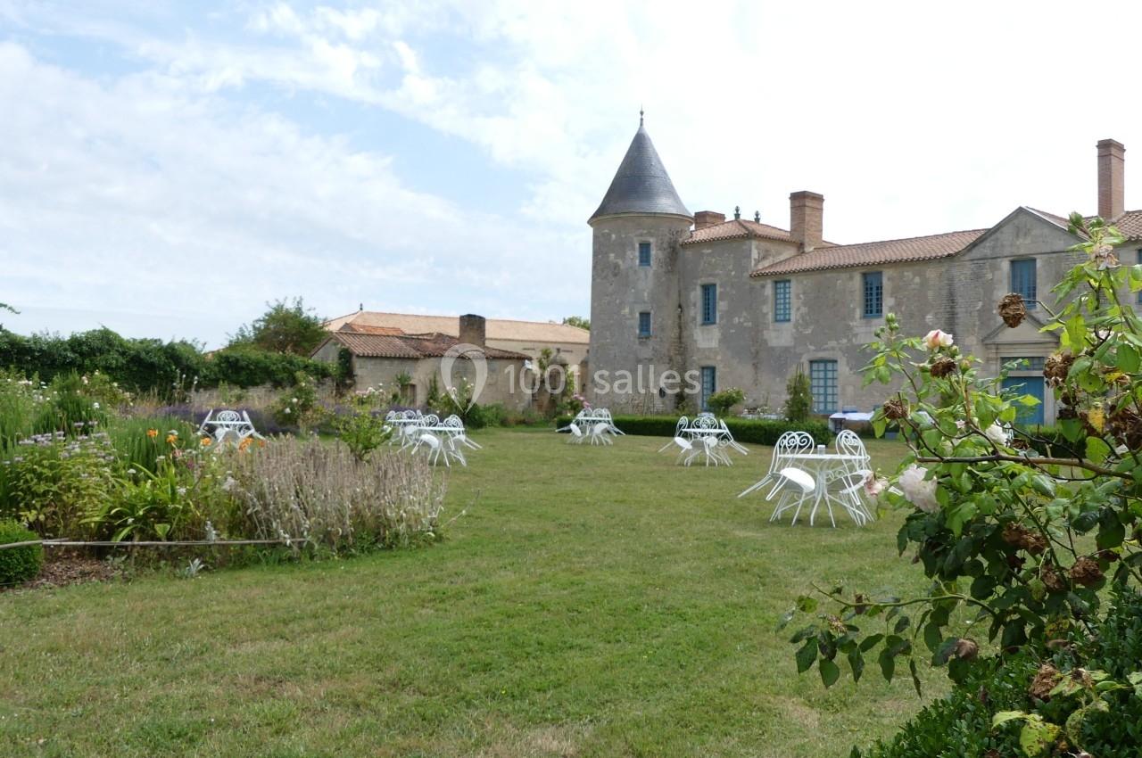 Location salle Sainte-Gemme-la-Plaine (Vendée) - Château de la Chevallerie #43 Jardin avec tables et chaises blanches devant un bâtiment ancien en pierre avec une tour et un toit en tuiles.