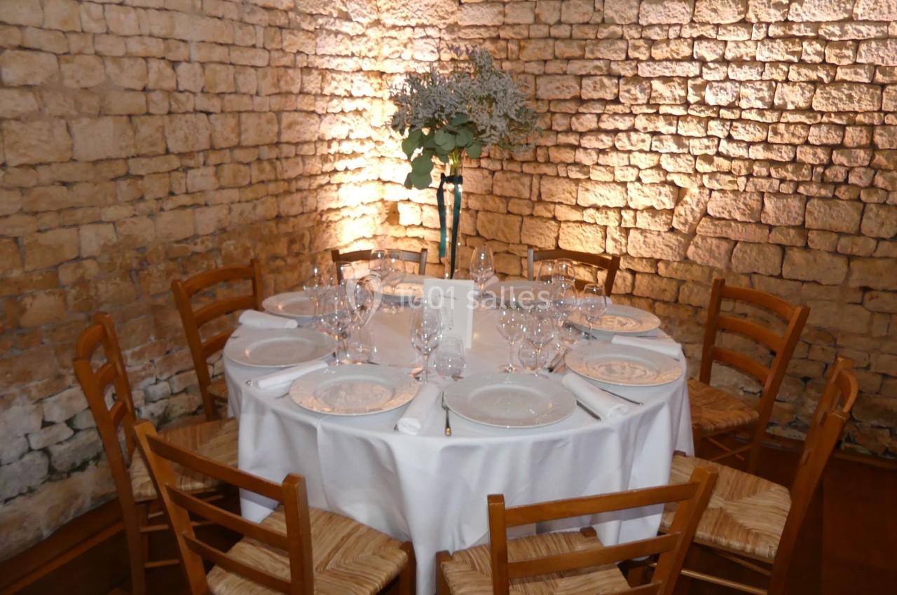 Location salle Sainte-Gemme-la-Plaine (Vendée) - Château de la Chevallerie #20 Table ronde dressée avec nappes blanches, vaisselle et bouquet central, entourée de chaises en bois, devant un mur en pierre.