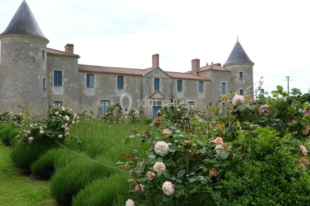 Location salle Sainte-Gemme-la-Plaine (Vendée) - Château de la Chevallerie #18 Château en pierre avec tours rondes, entouré de rosiers en fleurs et d'une pelouse verdoyante.