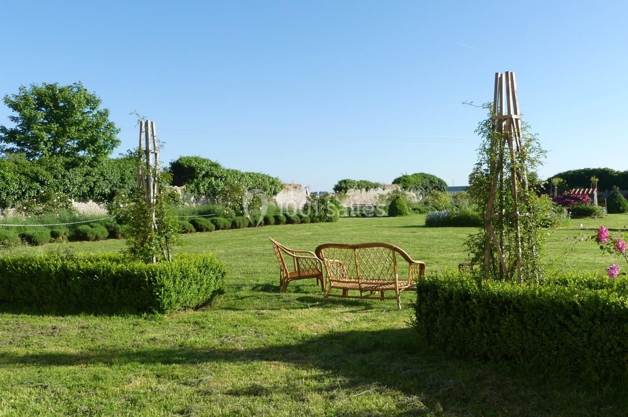 Location salle Sainte-Gemme-la-Plaine (Vendée) - Château de la Chevallerie #44 Mobilier en rotin disposé dans un jardin verdoyant entouré de haies et de structures végétales.