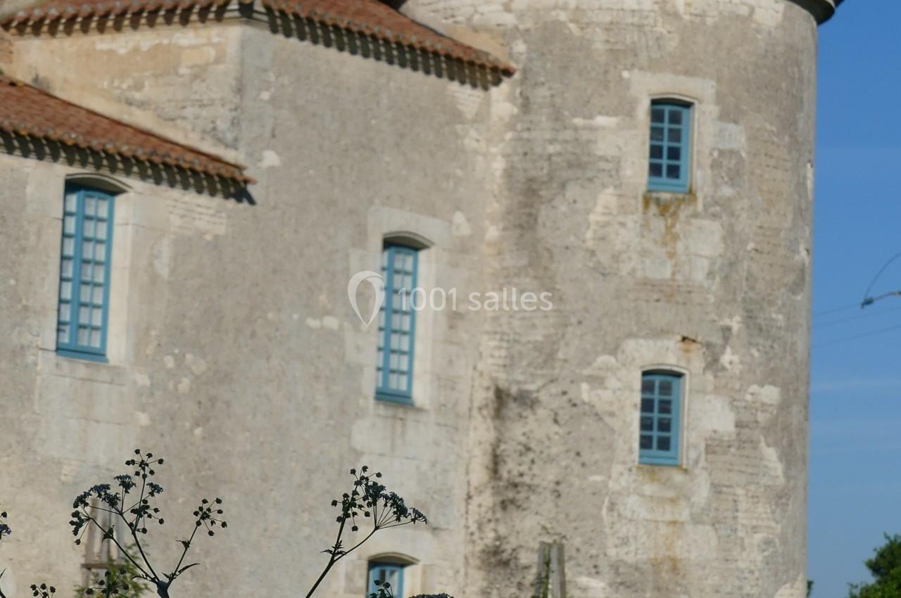 Location salle Sainte-Gemme-la-Plaine (Vendée) - Château de la Chevallerie #45 Tour en pierre d'un bâtiment ancien avec des fenêtres bleues, entourée de verdure et de mobilier de jardin en rotin.