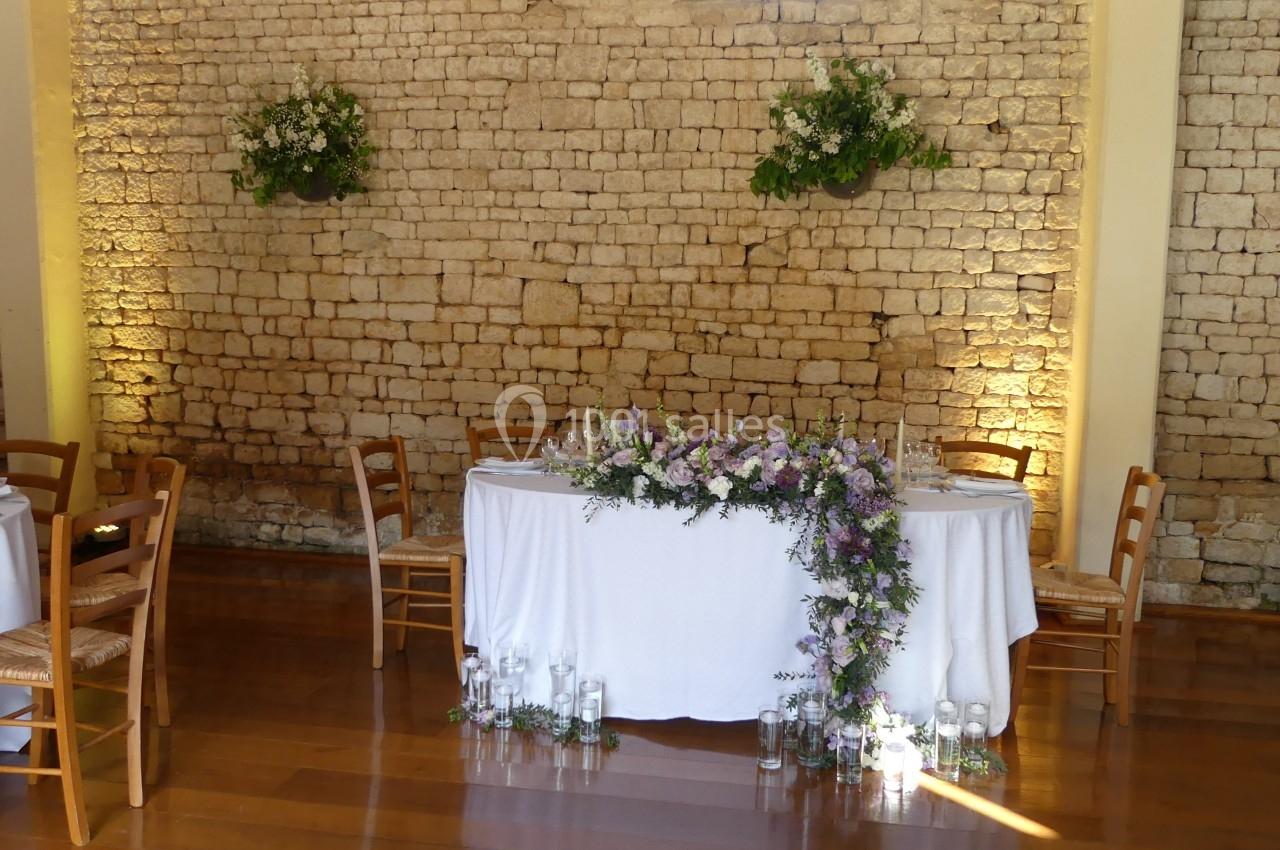 Location salle Sainte-Gemme-la-Plaine (Vendée) - Château de la Chevallerie #7 Table décorée pour un événement, ornée de fleurs violettes et blanches, devant un mur en pierre éclairé.