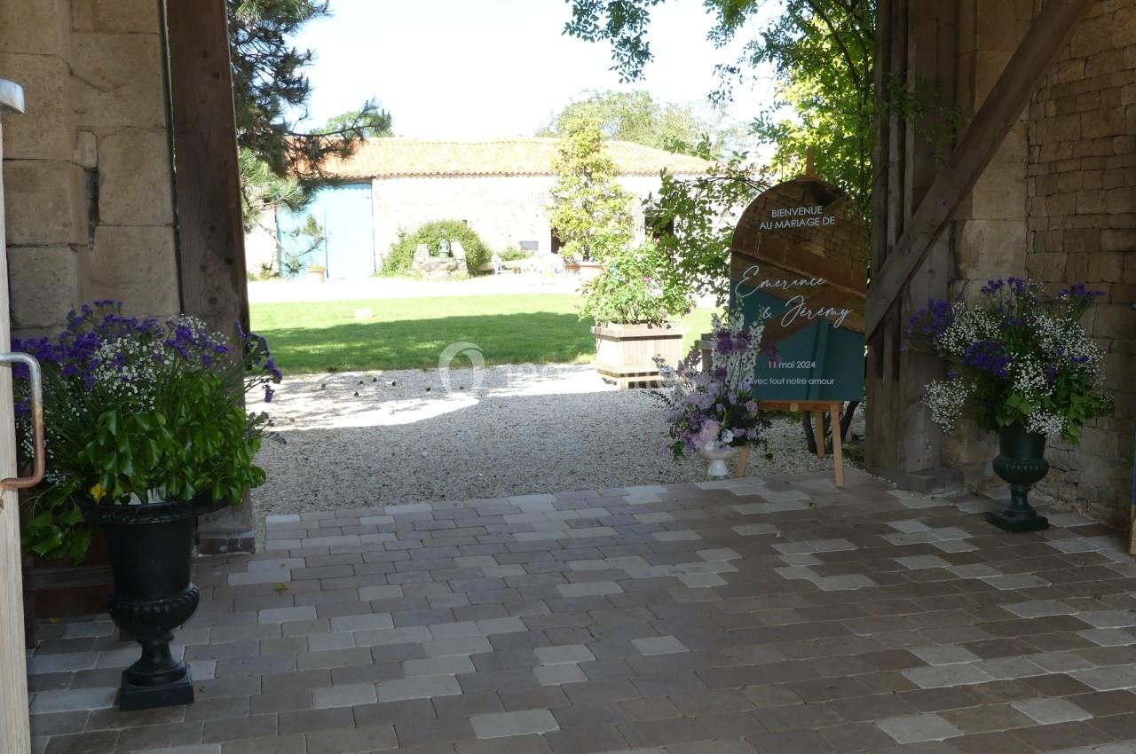 Location salle Sainte-Gemme-la-Plaine (Vendée) - Château de la Chevallerie #4 Entrée pavée d'une cour avec des fleurs en pots, une enseigne en bois et un jardin en arrière-plan.