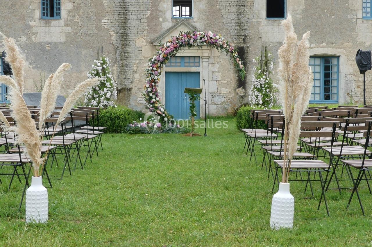 Location salle Sainte-Gemme-la-Plaine (Vendée) - Château de la Chevallerie #46 Allée centrale d'une cérémonie en plein air, décorée d'herbes de la pampa et d'une arche fleurie devant un bâtiment ancien.