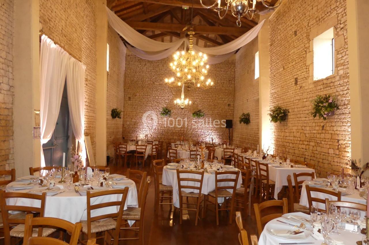 Location salle Sainte-Gemme-la-Plaine (Vendée) - Château de la Chevallerie #6 Salle de réception en pierre avec tables rondes dressées, chaises en bois, lustres suspendus et décorations florales.