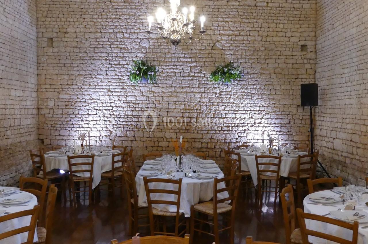 Location salle Sainte-Gemme-la-Plaine (Vendée) - Château de la Chevallerie #48 Salle en pierre avec tables rondes dressées, chaises en bois, chandelier central et décorations végétales murales.