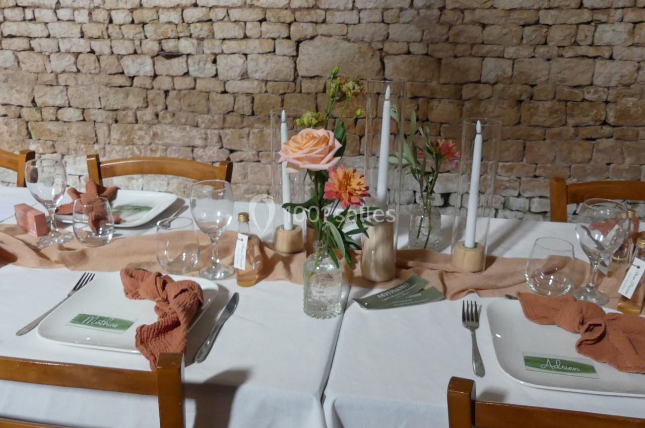 Location salle Sainte-Gemme-la-Plaine (Vendée) - Château de la Chevallerie #5 Table dressée avec nappes blanches, serviettes orange, fleurs, bougies et vaisselle devant un mur en pierre.