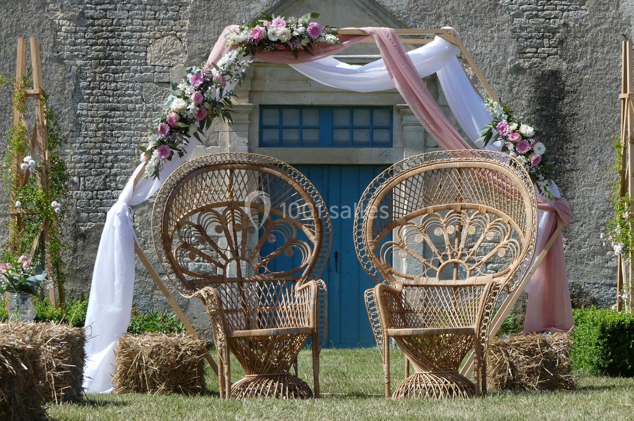 arche hexagonale Deux fauteuils en rotin devant une arche décorée de fleurs et de tissus, installée près d'une porte bleue sur un mur en…