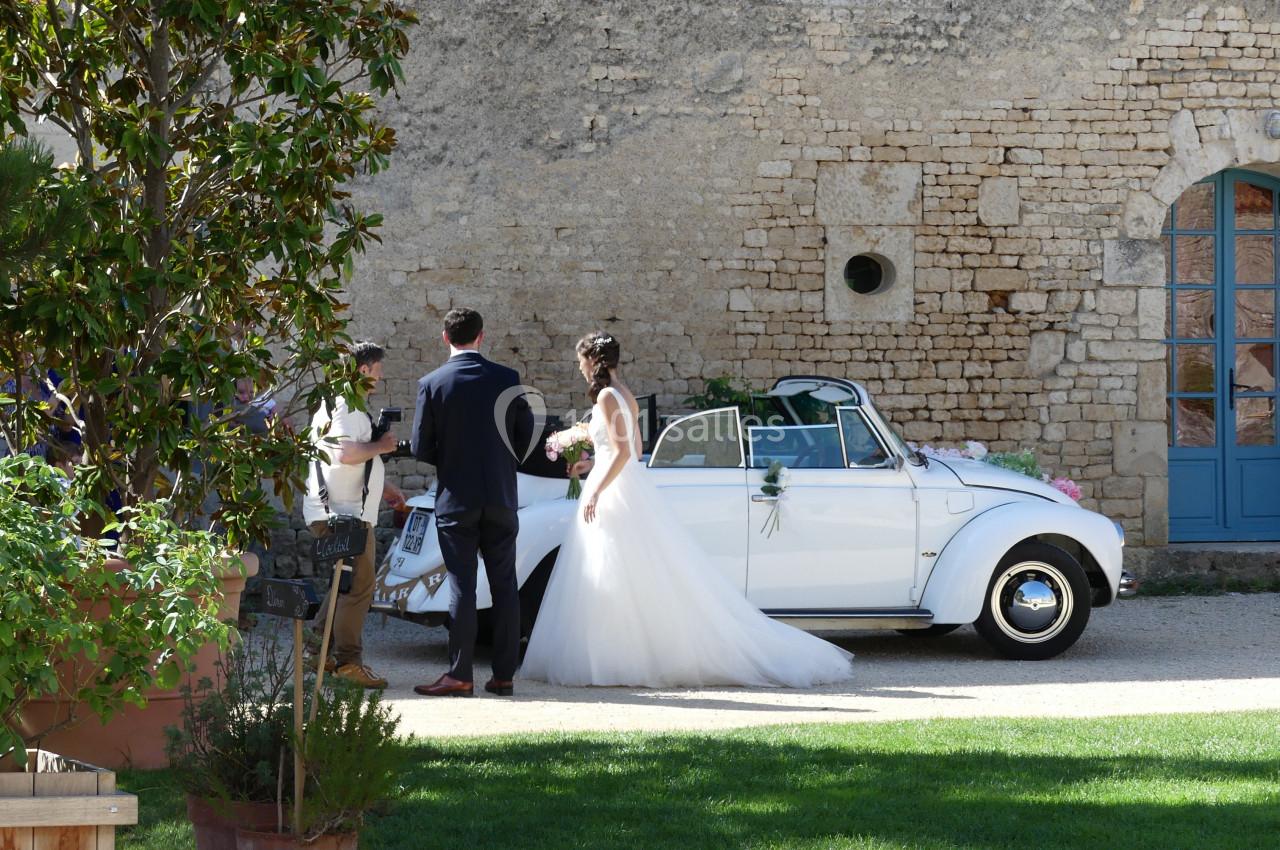 Arrivée des mariés Un couple de mariés se tient près d'une voiture blanche décapotable décorée, devant un mur en pierre.