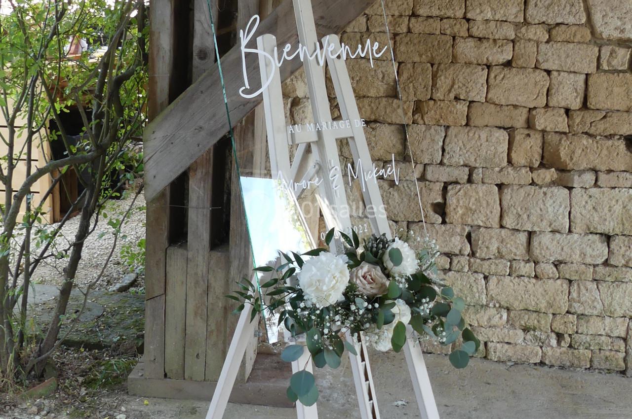 bienvenue à La Chevallerie Panneau de bienvenue pour un mariage, en verre transparent, décoré de fleurs blanches et vertes, posé sur un chevalet.