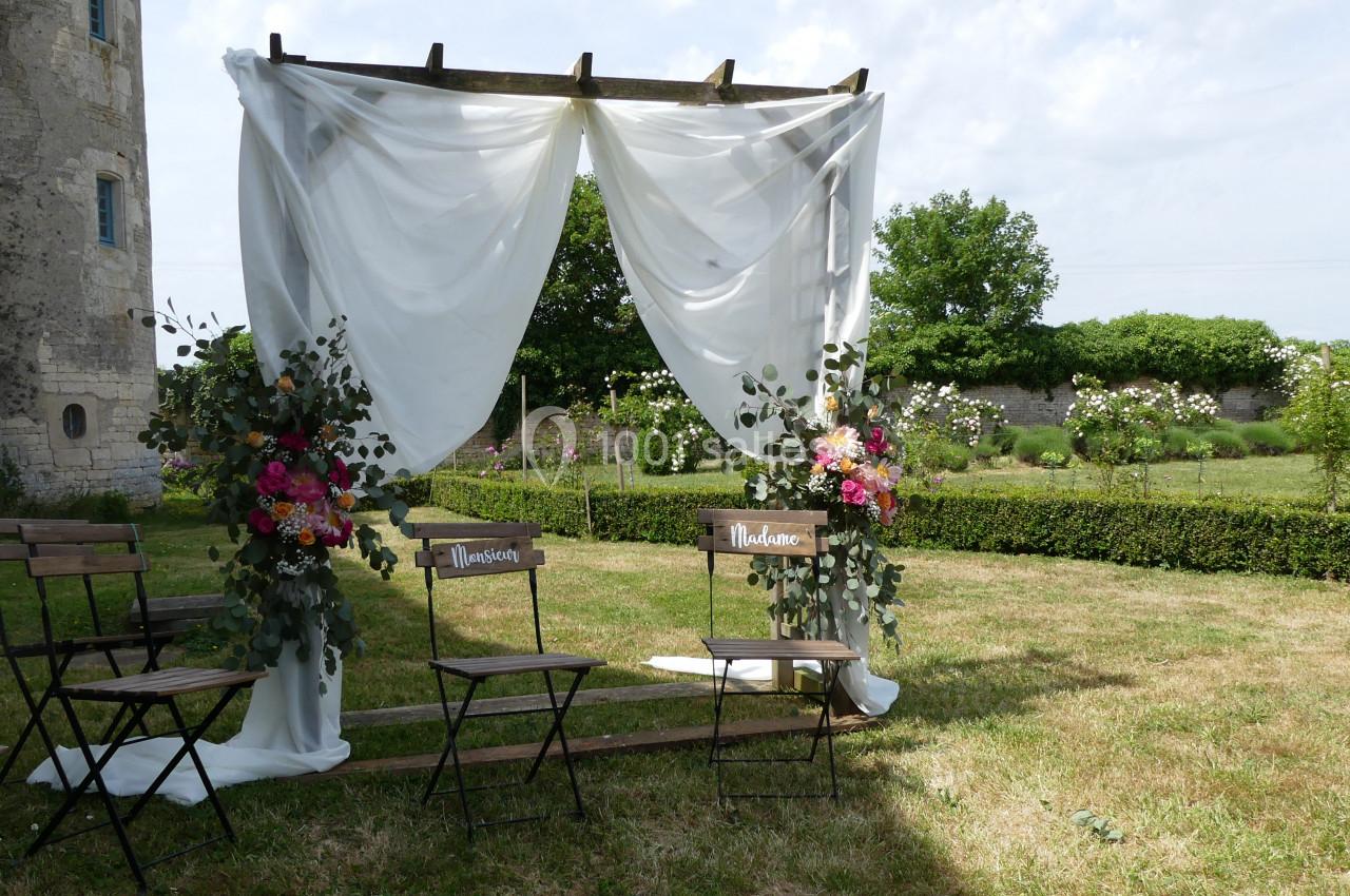 Cérémonie à l'ombre du château Arche de mariage en bois décorée de voilages blancs et de fleurs colorées, avec deux chaises sur une pelouse.