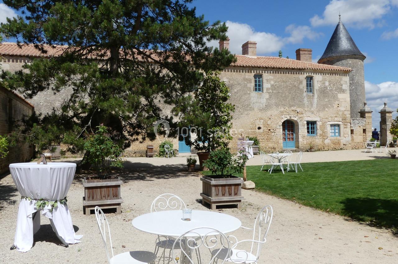 Cour du Pavillon pour votre cocktail Cour d'un château avec mobilier de jardin blanc, table dressée et pelouse, sous un ciel partiellement nuageux.