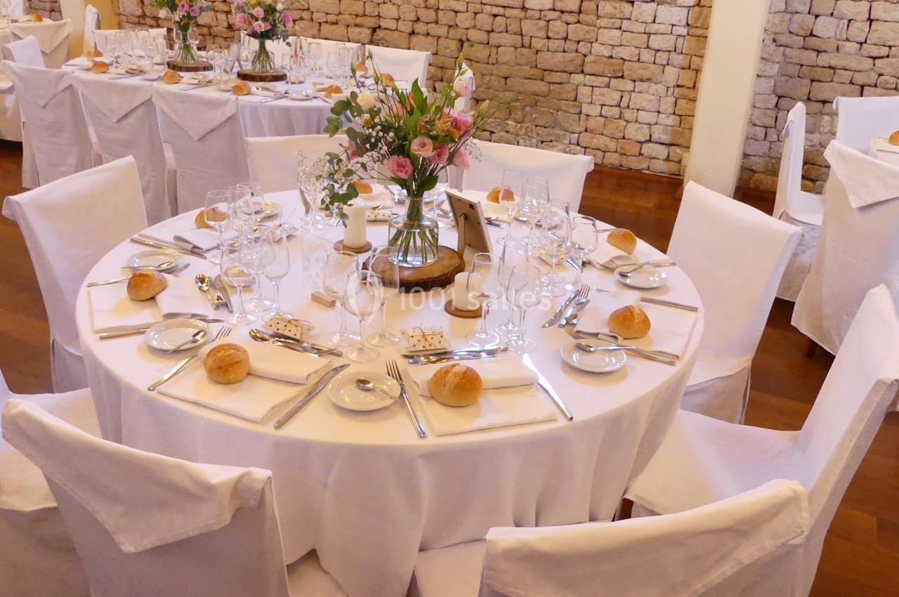 Des fleurs, des fleurs, mais de saison ! Table ronde élégamment dressée avec nappes blanches, vaisselle, fleurs au centre et chaises couvertes.