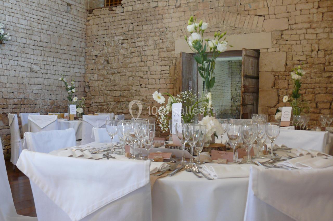Jolie salle ! Table de réception élégamment dressée avec nappes blanches, verres, couverts et décoration florale dans une salle en pierre.