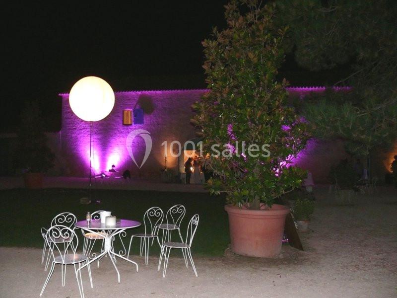 Nuit en couleur Cour extérieure éclairée de nuit avec des chaises blanches, une table et un bâtiment illuminé en arrière-plan.