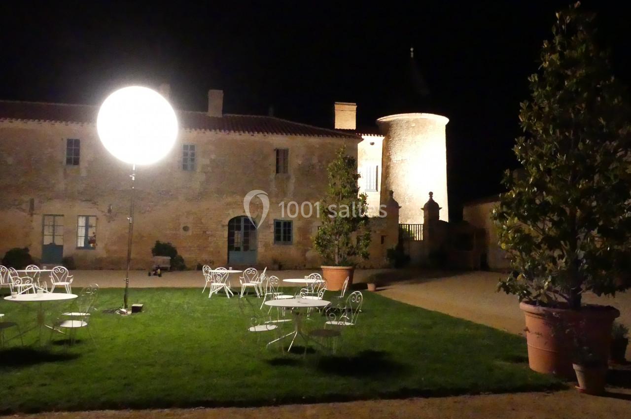 Pleine lune ! Cour éclairée la nuit avec des tables et chaises en fer forgé sur une pelouse, devant un bâtiment en pierre.