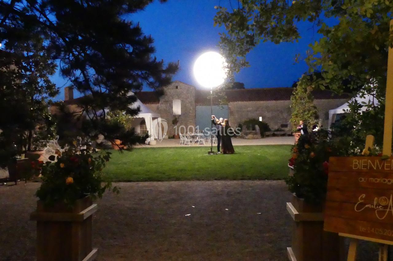 Romantique Un couple danse sous une lumière ronde dans un jardin éclairé, entouré de bâtiments en pierre au crépuscule.