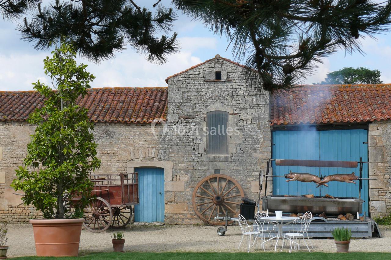 rotisserie en plein air Cour d'une ferme avec murs en pierre, charrette ancienne, table en fer forgé et rôtissoire en fonctionnement.