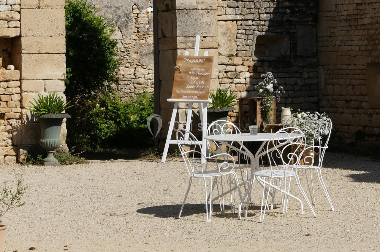 salon de jardin Table et chaises blanches en fer forgé disposées sur une terrasse en gravier, entourées de plantes et de murs en pierre.