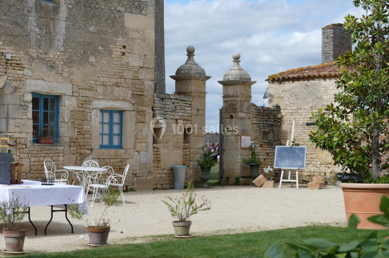 tables et chaises en fer forgées Cour d'une bâtisse en pierre avec mobilier de jardin, tableau noir et plantes en pot, sous un ciel partiellement nuageux.