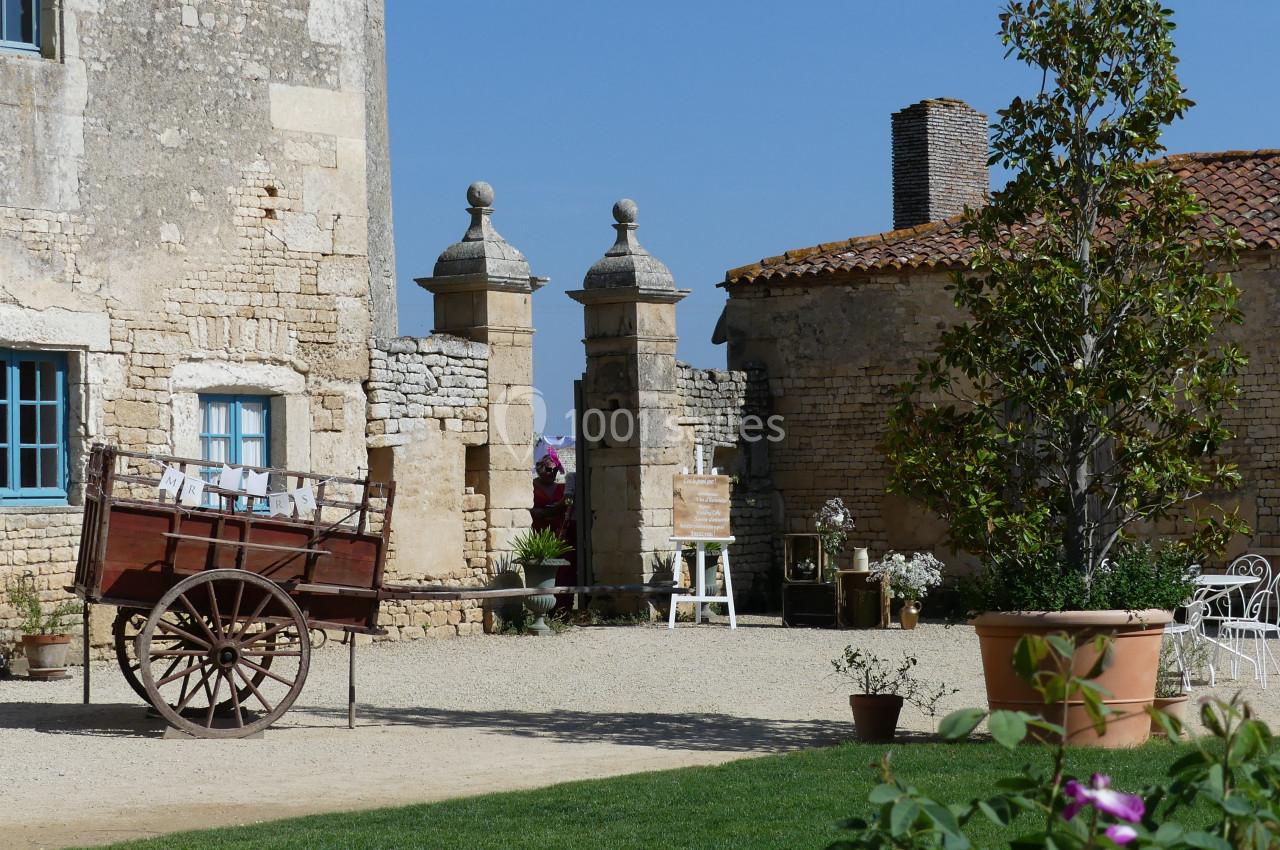 voiture ancienne Cour d'une bâtisse en pierre avec une charrette en bois, des plantes en pot et des meubles de jardin sous un ciel bleu.