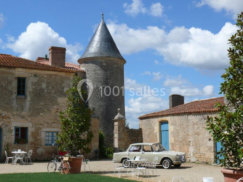Voiture des mariés Cour d'une bâtisse en pierre avec une tour, des tables en fer forgé, et une voiture ancienne stationnée.
