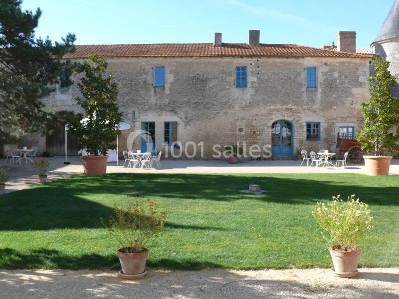 Votre maison pendant 4 jours ! Cour d'une maison en pierre avec pelouse, tables et chaises extérieures, entourée de pots de fleurs et d'arbres.