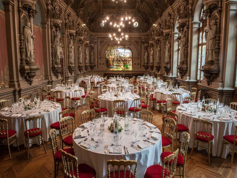 Salle de réception élégante avec tables rondes dressées, chaises rouges et décorations sculptées sur les murs.