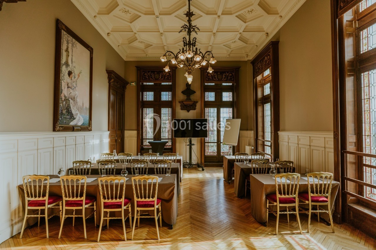 Salle élégante avec parquet, tables dressées, chaises dorées, lustre central et grandes fenêtres ornées de vitraux.