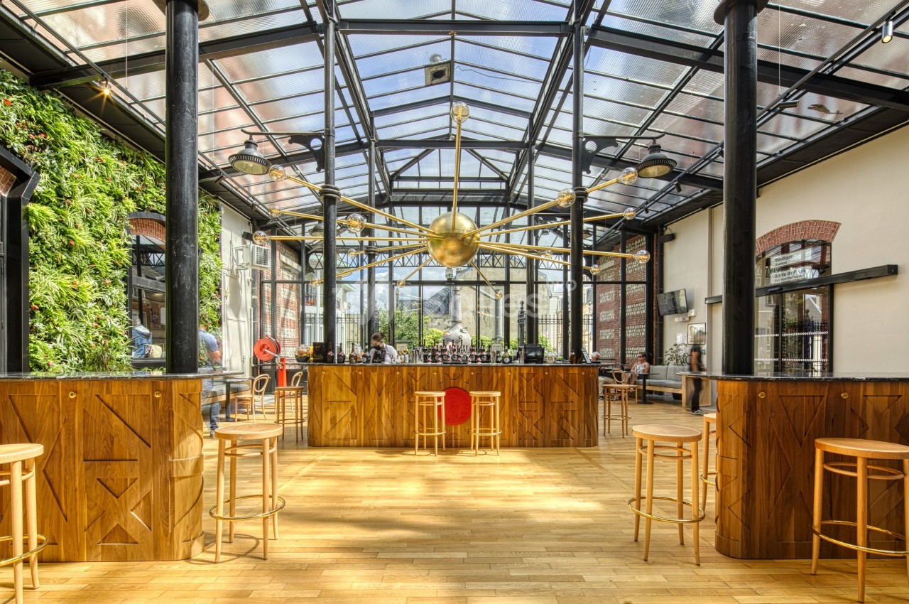 Intérieur d'un bar lumineux avec verrière, comptoir central en bois, tabourets et mur végétal sur le côté.